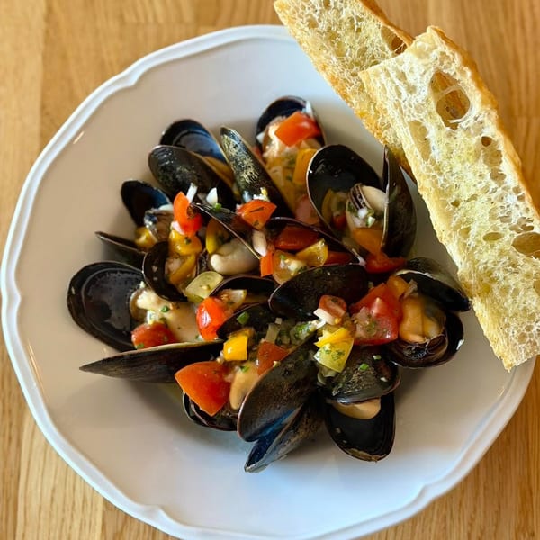 Steamed mussels topped with diced tomatoes and peppers, served with toasted bread on a white plate.