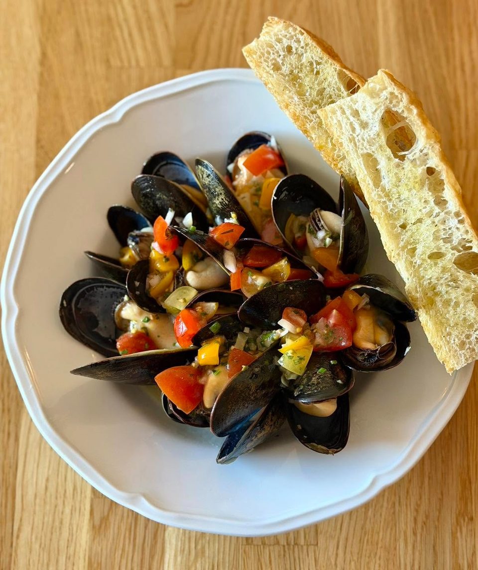 Steamed mussels topped with diced tomatoes and peppers, served with toasted bread on a white plate.
