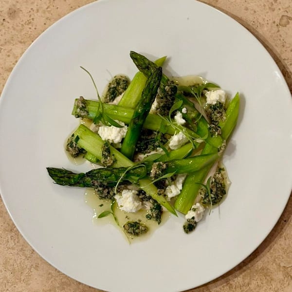 Bright green asparagus spears with crumbled white cheese, pesto, and microgreens on a white plate.