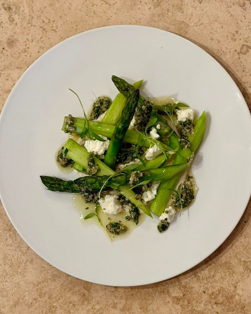 Bright green asparagus spears with crumbled white cheese, pesto, and microgreens on a white plate.