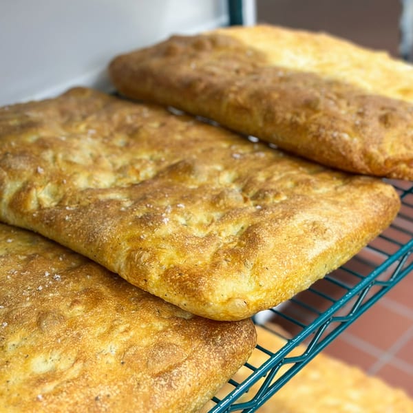 Golden-brown focaccia bread, sprinkled with salt and herbs, cooling on a green wire rack.