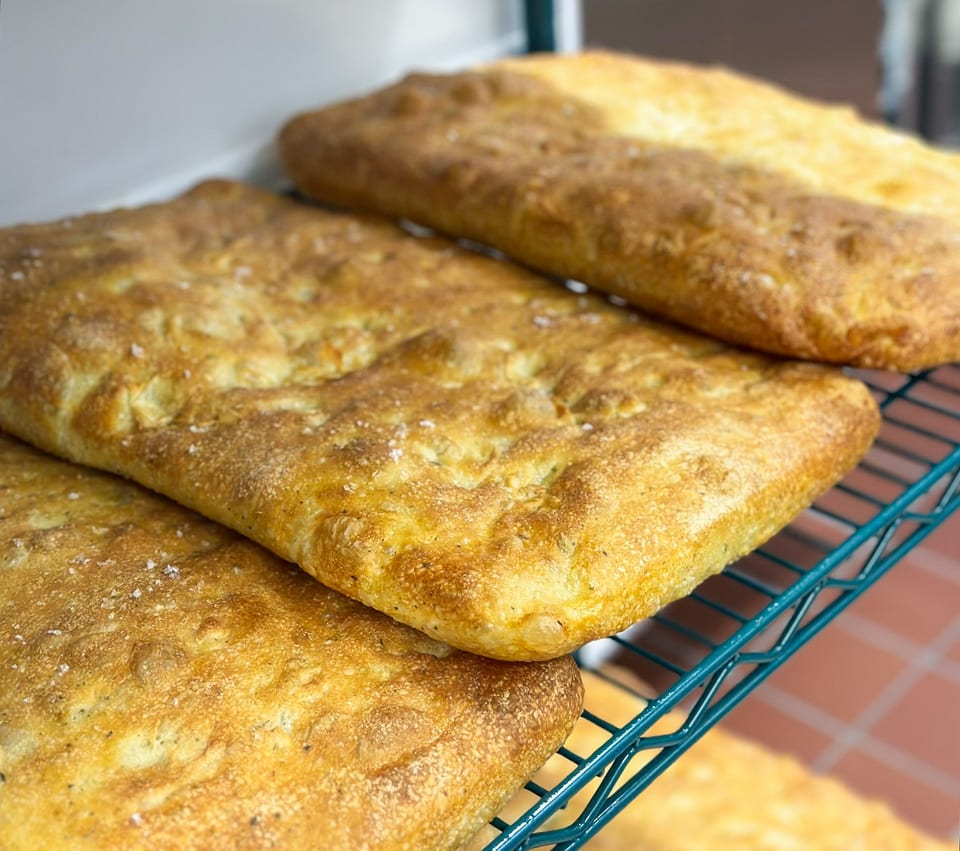 Golden-brown focaccia bread, sprinkled with salt and herbs, cooling on a green wire rack.