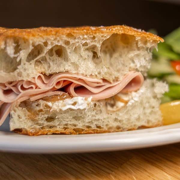 Close-up of a sandwich with ham, cheese, and caramelized onions on focaccia bread, with a side salad.