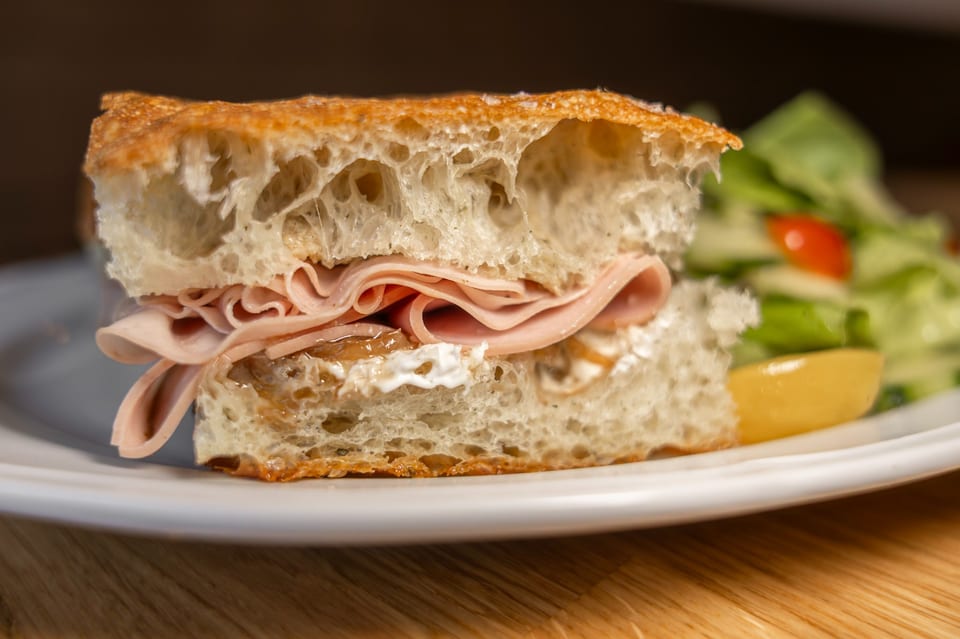 Close-up of a sandwich with ham, cheese, and caramelized onions on focaccia bread, with a side salad.