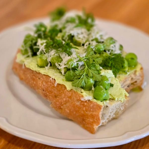 Toasted baguette with green pea spread, whole peas, fresh herbs, and grated cheese on a white plate.