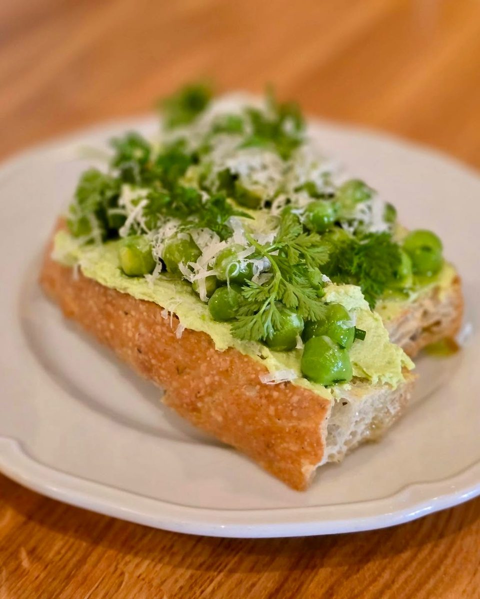 Toasted baguette with green pea spread, whole peas, fresh herbs, and grated cheese on a white plate.