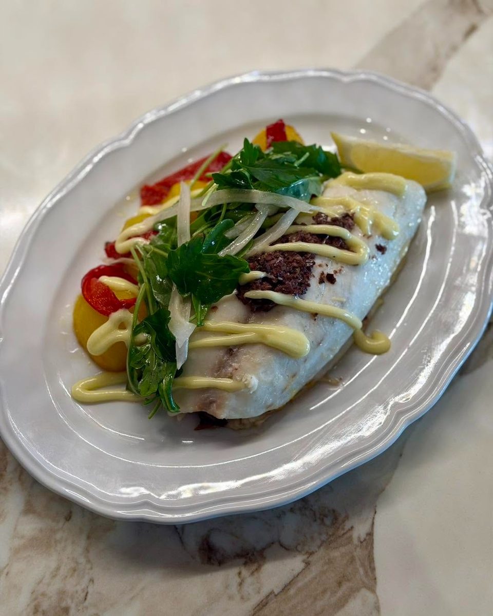 White fish fillet with olive tapenade, mayonnaise drizzle, herbs, bell peppers, and a lemon wedge.