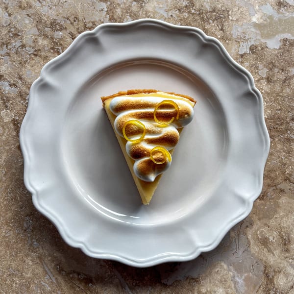 A slice of lemon meringue pie with toasted meringue and candied lemon peel on a white plate.