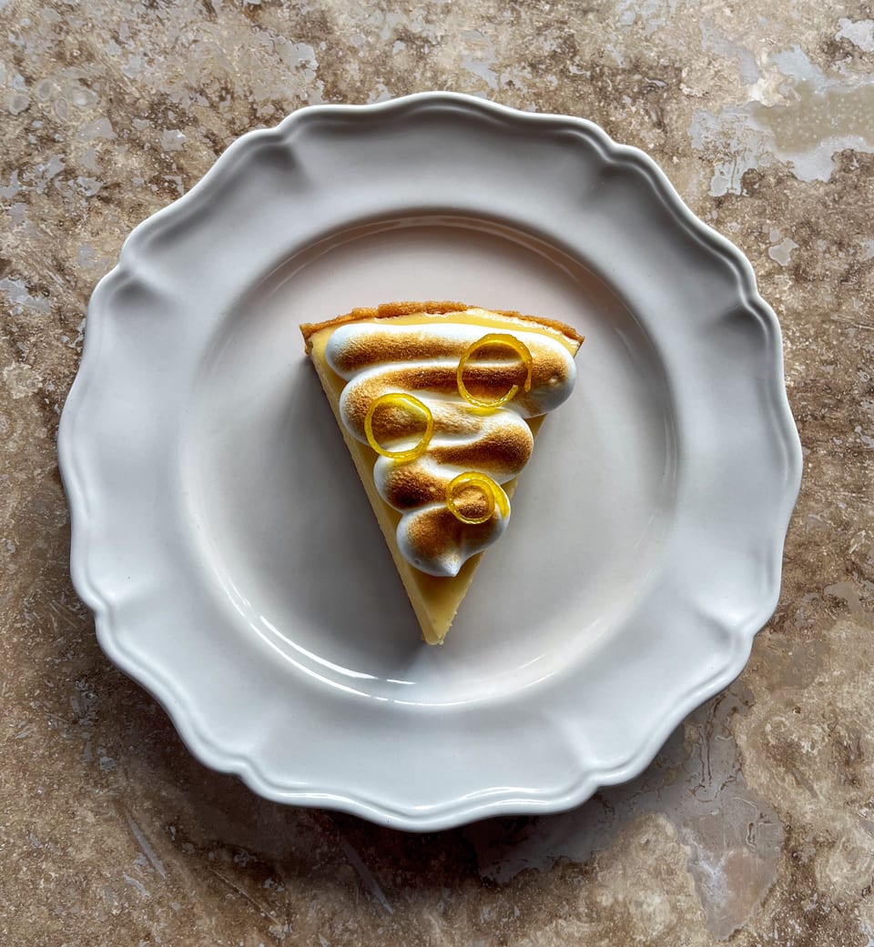 A slice of lemon meringue pie with toasted meringue and candied lemon peel on a white plate.