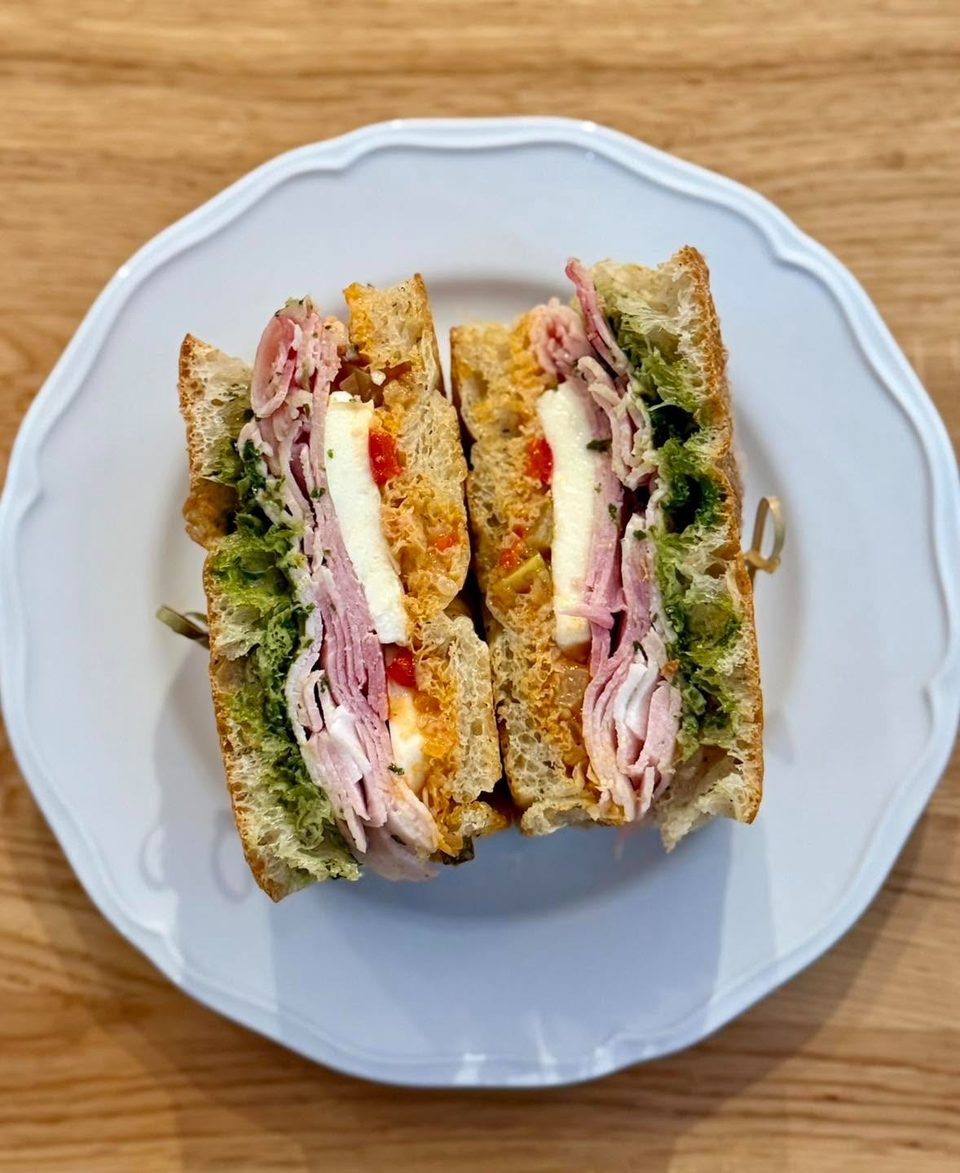 A sliced sandwich with ham, cheese, pesto, and red peppers on a white plate.