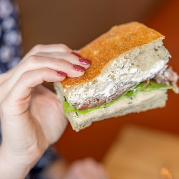 A hand with red nail polish holds a focaccia sandwich filled with meat, lettuce, and white sauce.