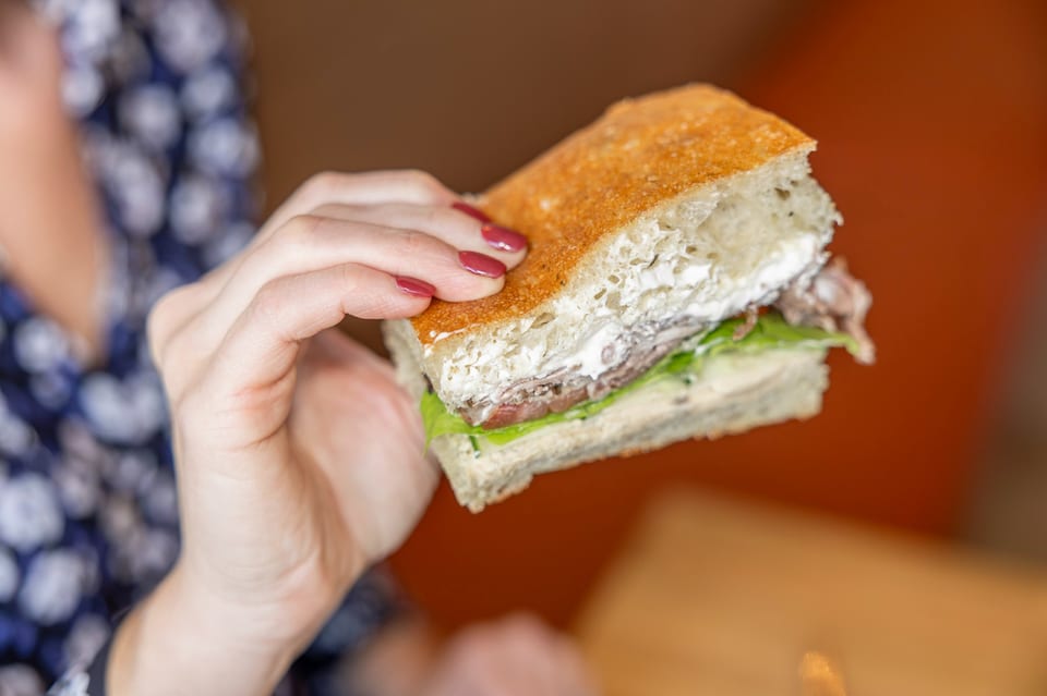 A hand with red nail polish holds a focaccia sandwich filled with meat, lettuce, and white sauce.